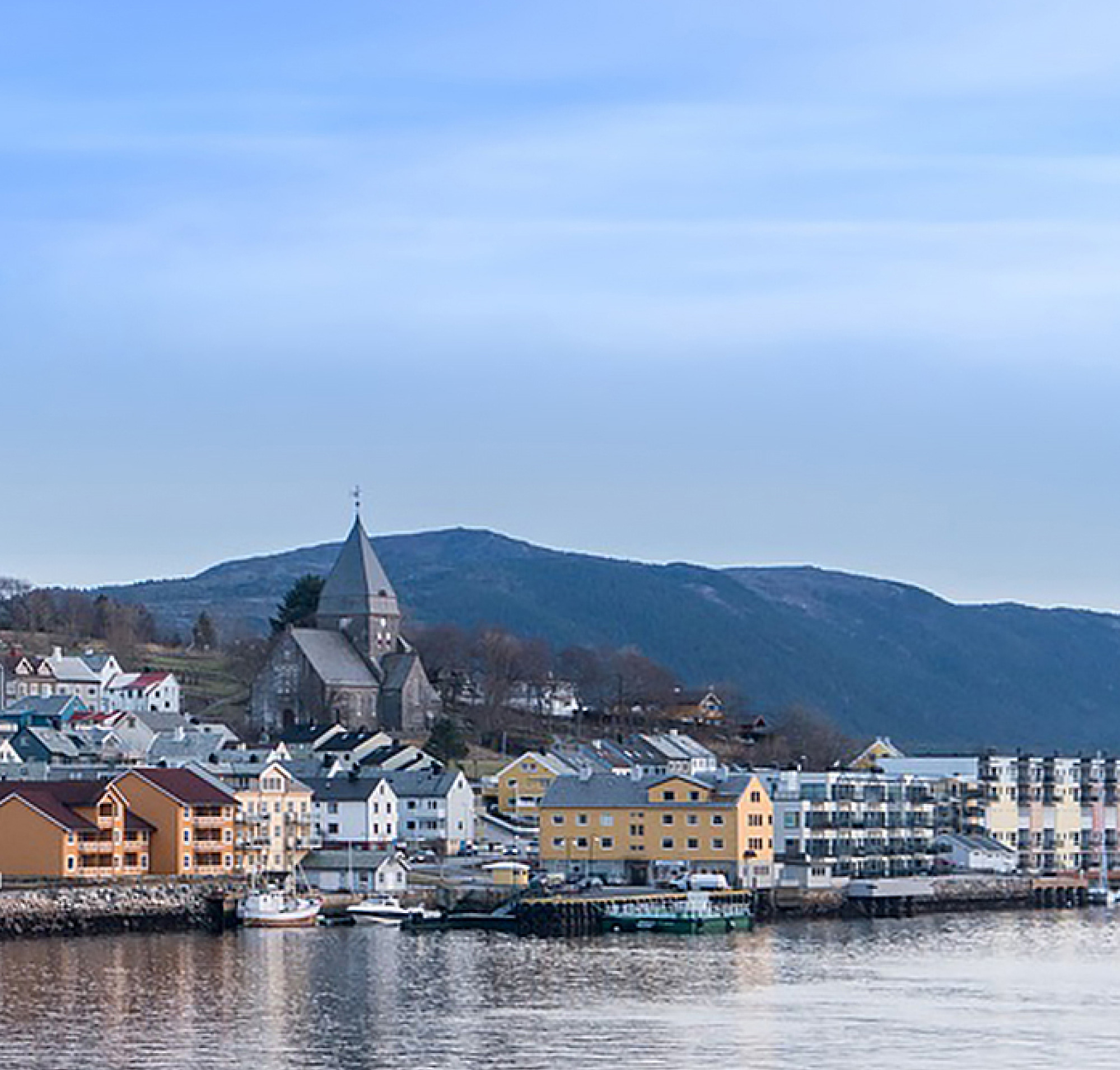 Vista panorâmica de uma cidade costeira norueguesa com edifícios coloridos e uma igreja proeminente, tendo como pano de fundo as colinas 