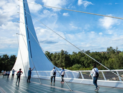 Alcune persone che passeggiano su un moderno ponte sospeso con strutture bianche a forma di vela, circondate da alberi
