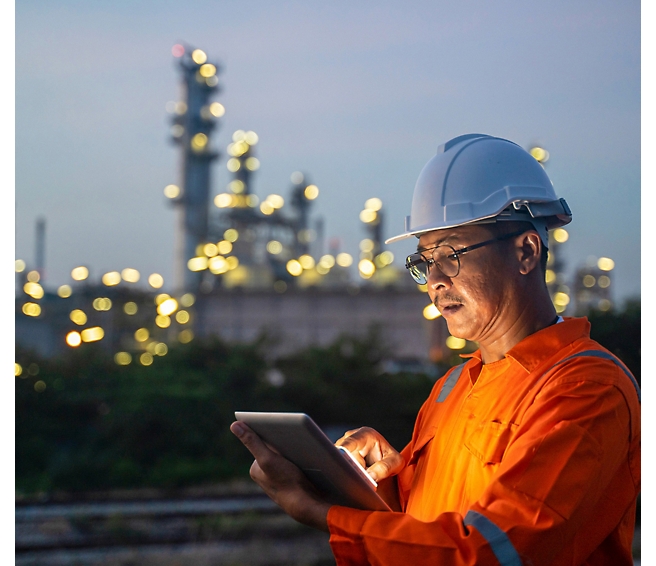 Ingeniero con un mono naranja y un casco que usa una tableta, con una planta industrial iluminada al atardecer en segundo plano.