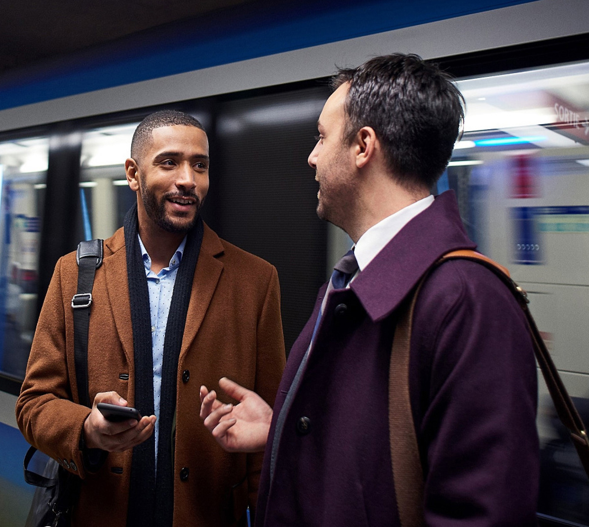 Two men conversing on a subway platform, with one holding a smartphone and a train blurred in motion behind them.