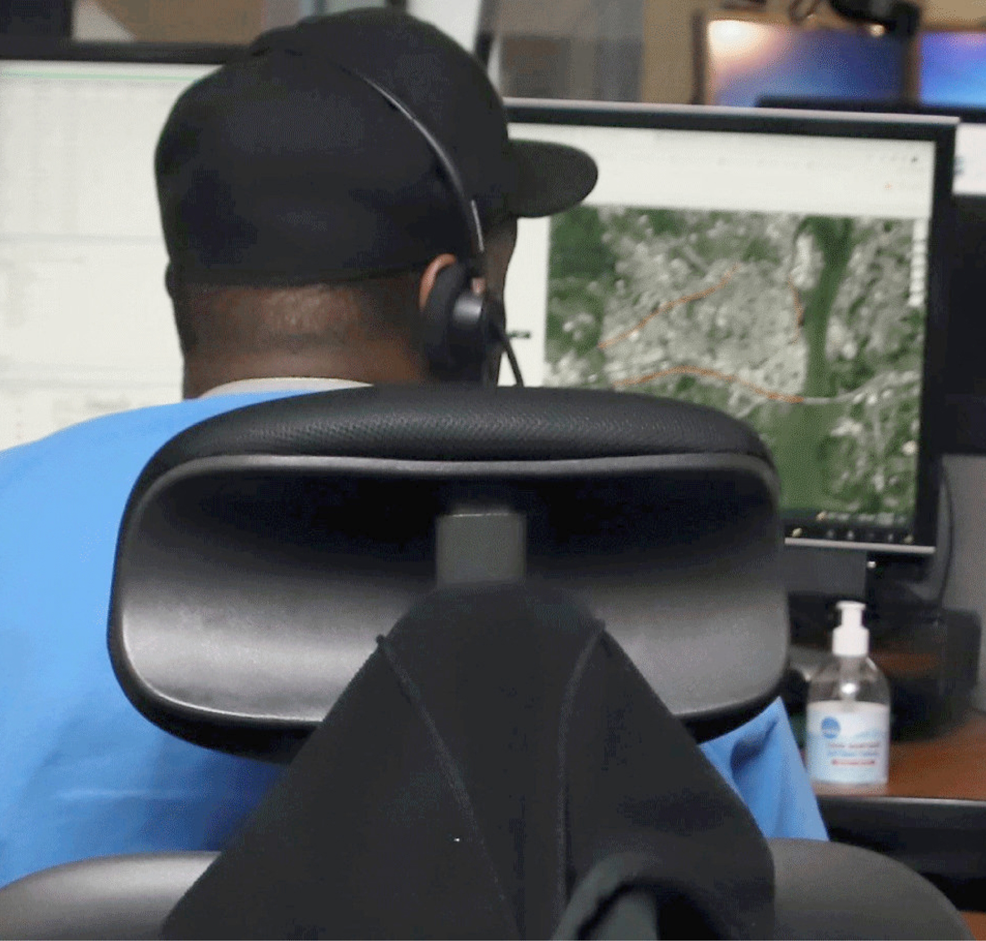 Man in orange shirt and black cap sitting at a desk, wearing headphones and looking at multiple computer screens.