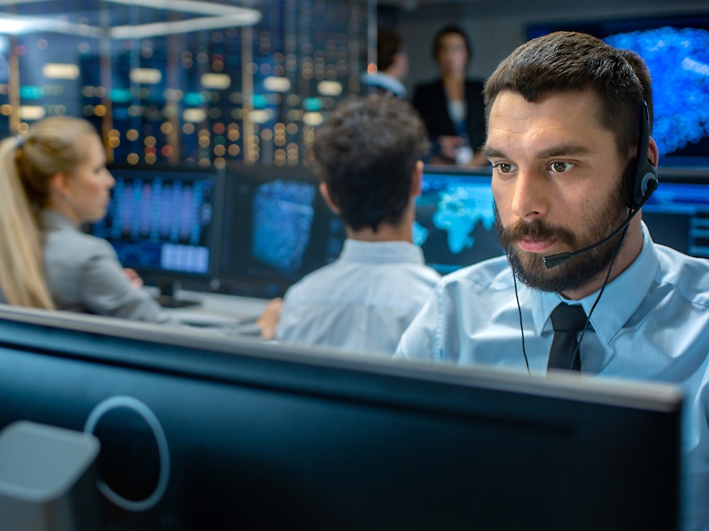 Seorang pria menggunakan headset bekerja menggunakan komputer di ruang kontrol yang ramai dengan rekan kerja dan layar besar menampilkan data