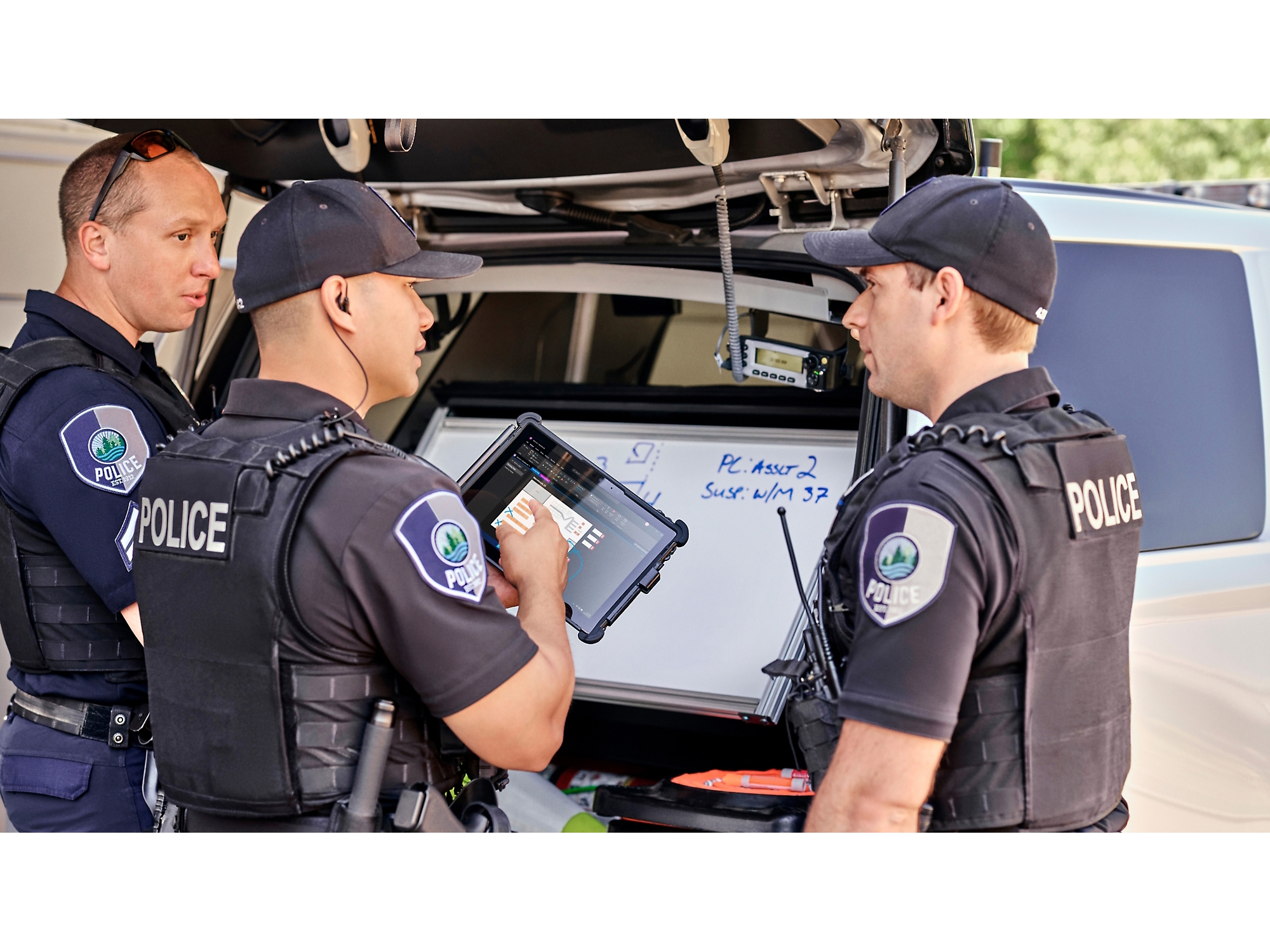 Drei Polizeibeamte prüfen Informationen auf einem digitalen Tablet neben ihrem Streifenwagen mit geöffnetem Kofferraum