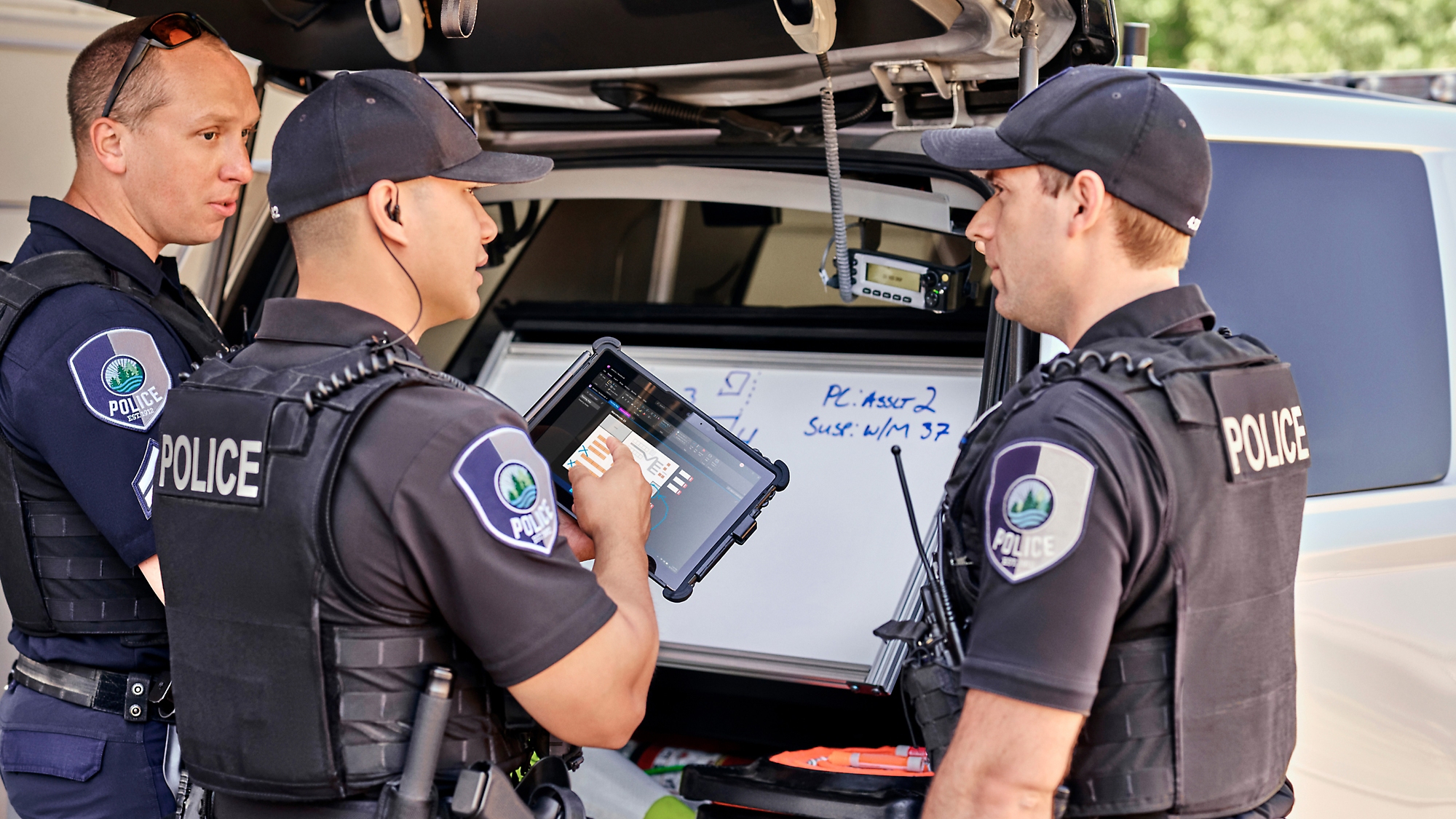 Three police officers review information on a digital tablet beside their patrol car with the trunk open