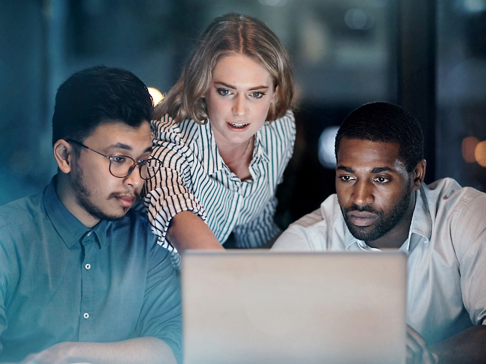 Três colegas, uma mulher e dois homens focam-se atentamente num ecrã de portátil num ambiente de escritório com iluminação fraca.