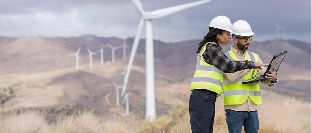 A woman in a yellow vest and white helmet standing in front of wind turbines.