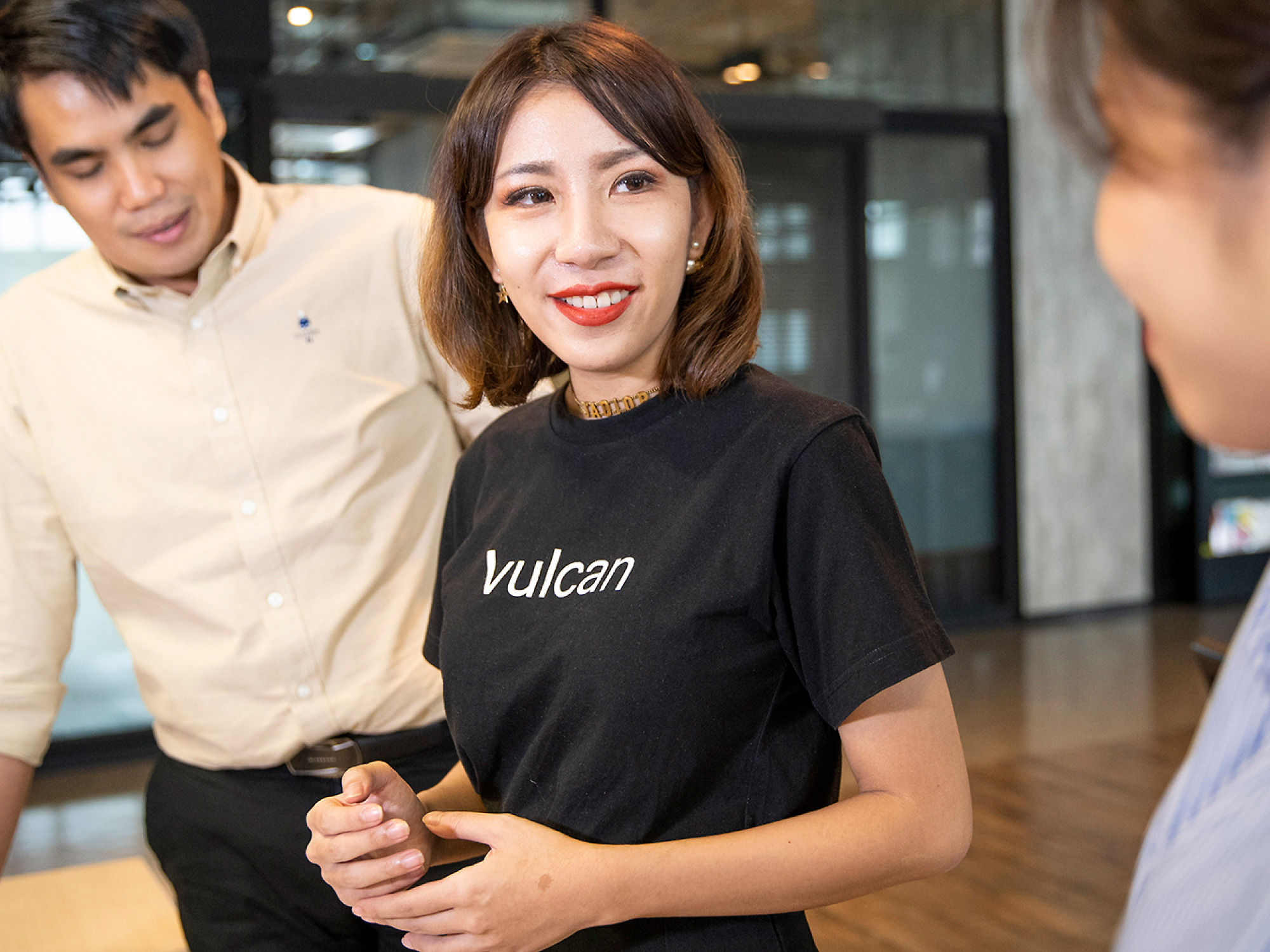 A young woman in black t-shirt labeled vulcan smiling at a coworker in a modern office setting, with another male colleague