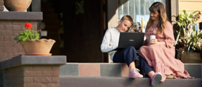 Dečak koji koristi laptop sedi na stepenicama trema pored žene sa pićem, u pratnji malog psa, u stambenom okruženju.
