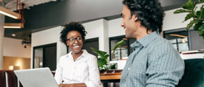 Two people are sitting and smiling at each other in an indoor sitting with a laptop on the table.