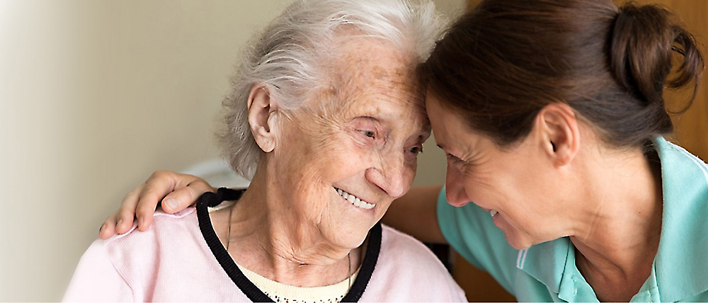 A caregiver smiling affectionately at an elderly woman who is also smiling, in a warm indoor setting.