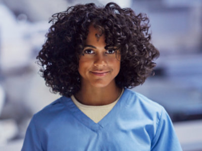 A person with curly hair wearing blue scrubs, standing in an indoor environment.