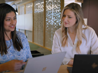 Two women sit at a table working on laptops in an office setting. They are engaged in a discussion.