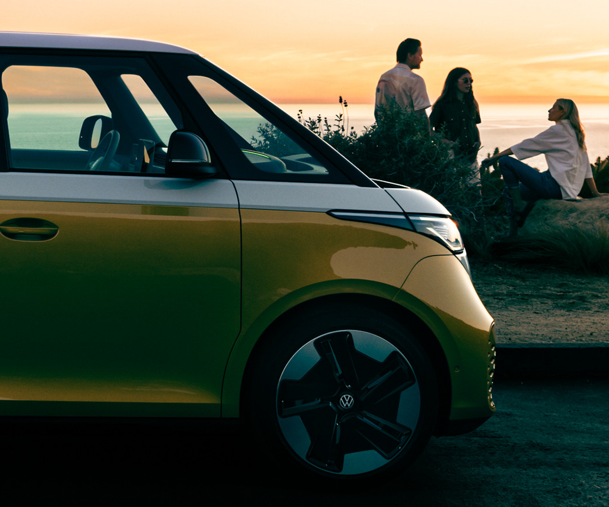 A yellow Volkswagen van parked on a coastal road at sunset with three people sitting and talking in the background.