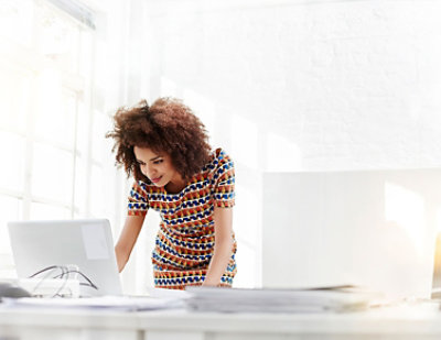 A person wearing a colorful dress stands at a white desk, leaning forward and looking at a laptop screen in a well-lit office.