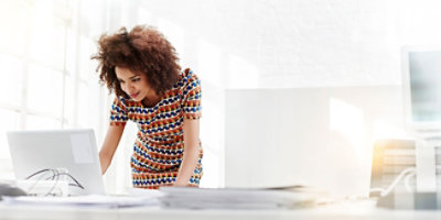 Osoba odevena u šarenu haljinu stoji za belim stolom u dobro osvetljenoj kancelariji, naginje se unapred i gleda u ekran laptopa.