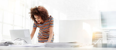A person wearing a colorful dress stands at a white desk, leaning forward and looking at a laptop screen in a well-lit office.