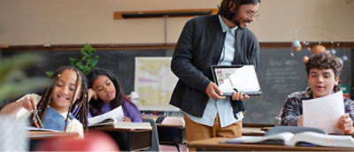 A teacher smiles as he holds a tablet, engaging with three students in a classroom.