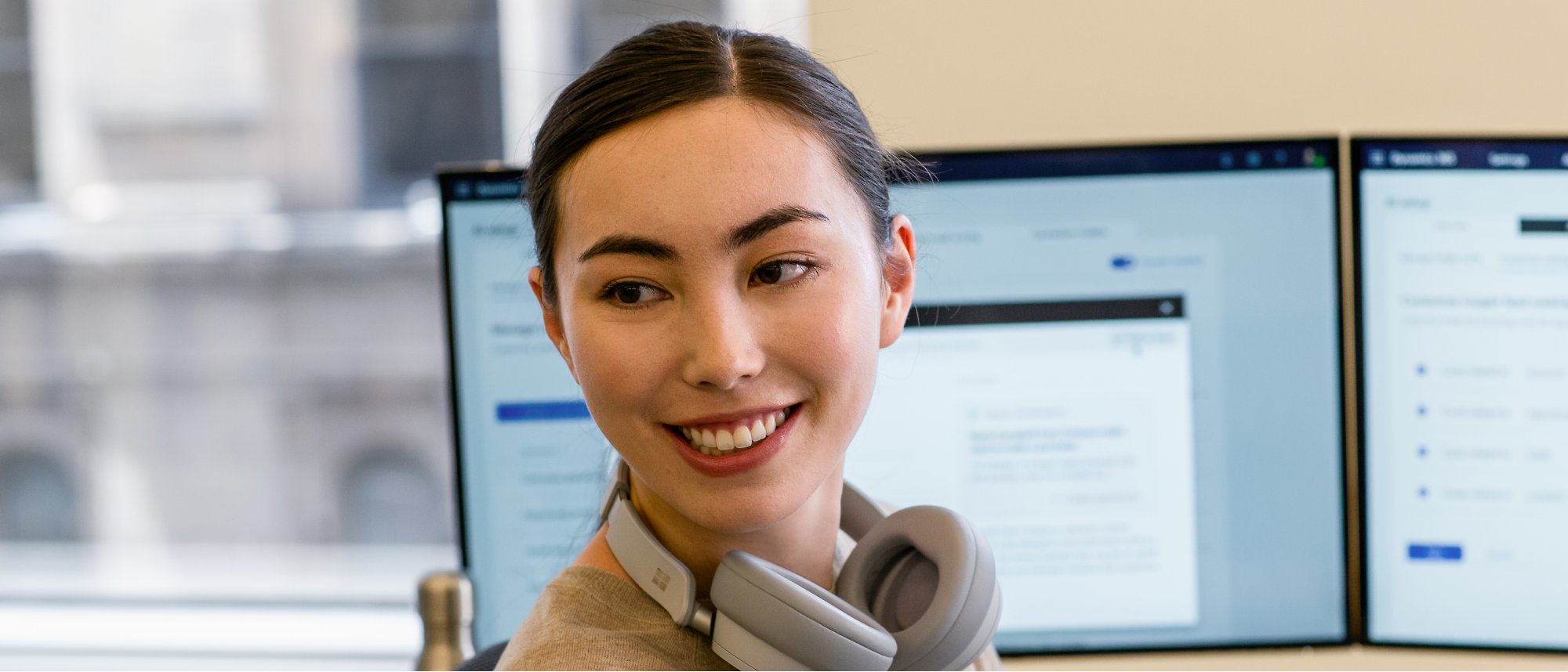Una mujer se pone el auricular sobre el cuello y sonríe y trabaja en un par de monitores.