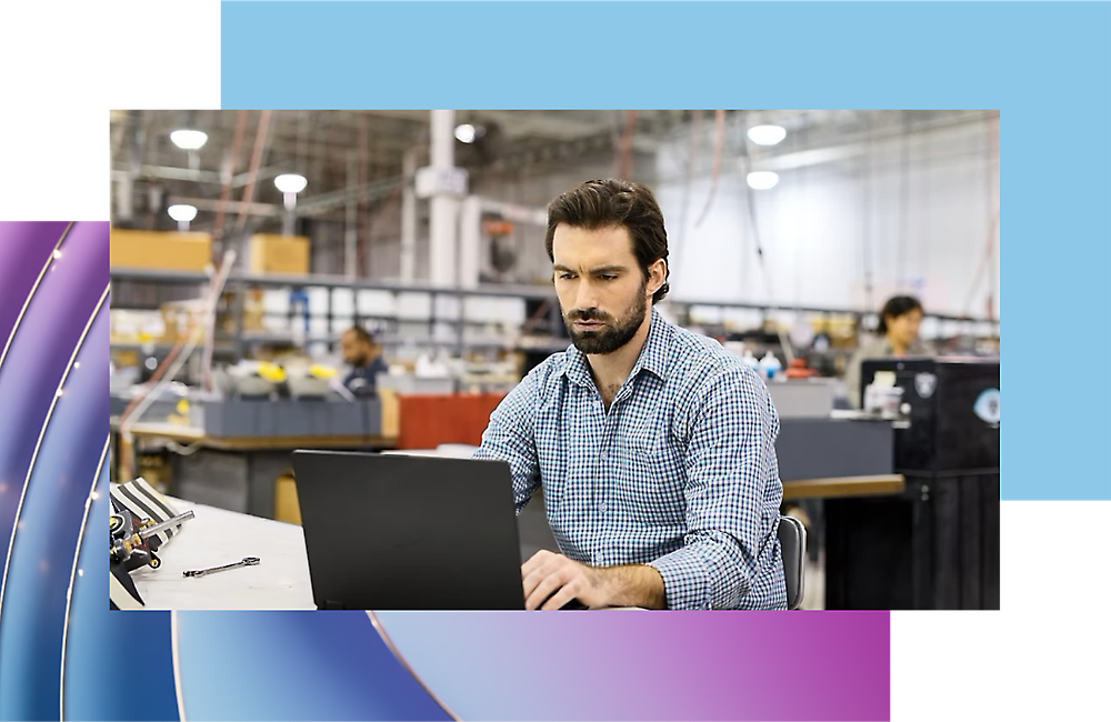 Ein Mann, der in einer Industrieumgebung an einem Laptop arbeitet. Im Hintergrund sind Maschinen und Arbeiter zu sehen.