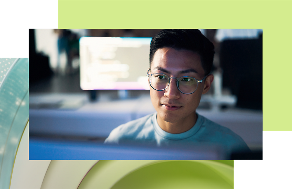眼鏡をかけてカメラを見ている若いアジア系の男性。背景でコンピューターの画面が光っている。