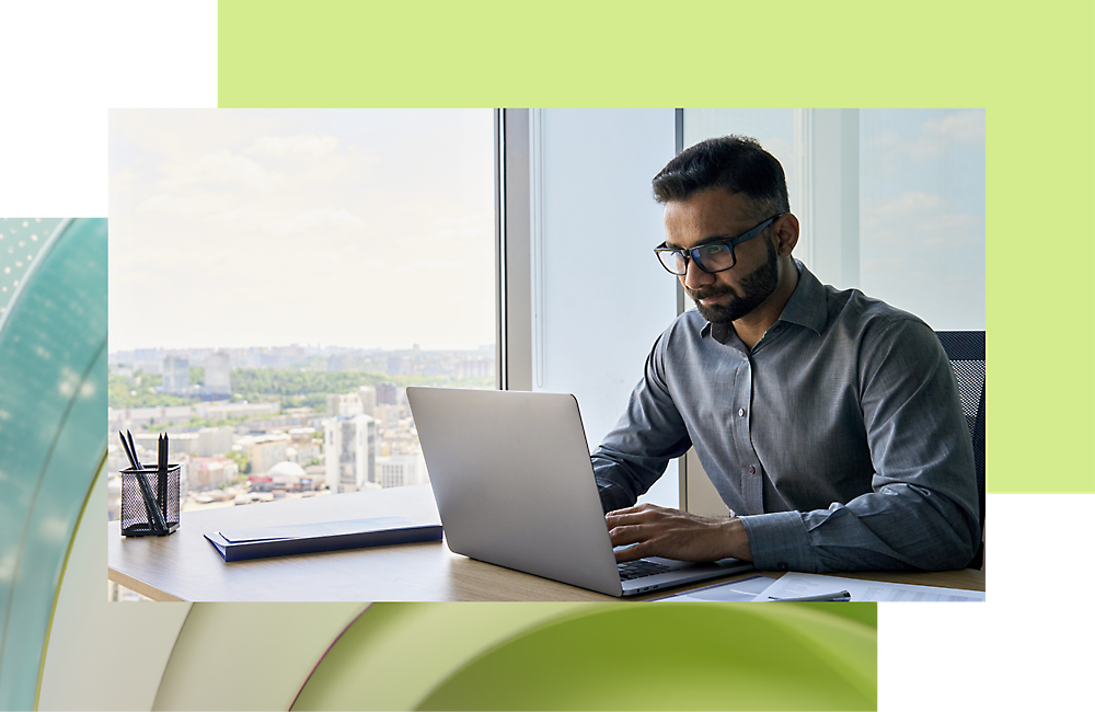 Mężczyzna w okularach, intensywnie pracujący na laptopie przy biurku przy oknie z widokiem na panoramę miasta.