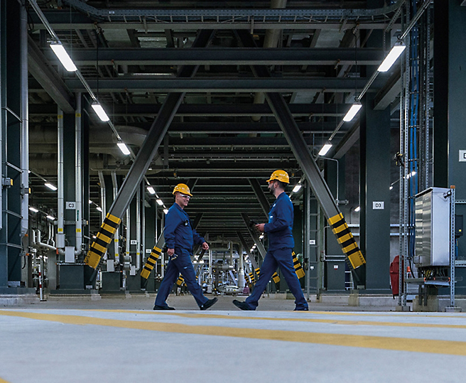 Dos trabajadores con cascos que caminan por una planta industrial.