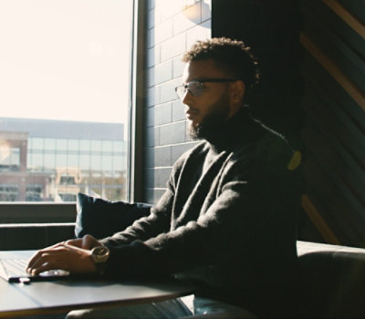 A man with a beard wearing glasses, sitting at a table using a laptop.