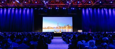 Large audience seated in a conference hall facing a stage with a city skyline displayed on a large screen.