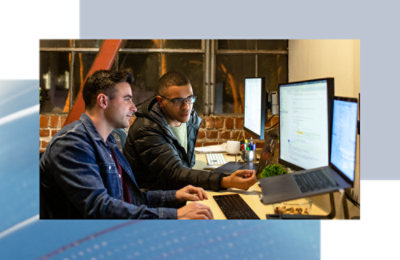 Two coworkers working side by side, reviewing code on multiple monitors in an office space.