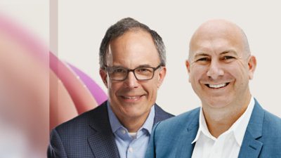 Twee mannen die glimlacht naar de camera. De een draagt ​​een bril en een colbert, terwijl de ander een lichtblauw jasje draagt.