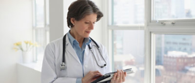 Clinician in a lab coat using a tablet near a window.