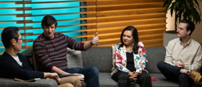 Group of people seated on a sofa in a modern office lounge engaged in a discussion, with notebooks and cups on a low table.