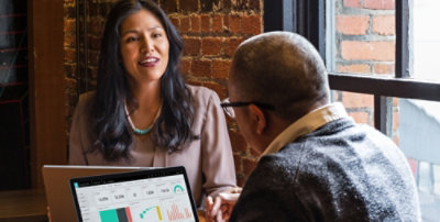 Two people reviewing Dynamics 365 sales data on laptop during a business meeting near a window