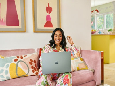 Seseorang duduk di sofa merah muda dan bekerja menggunakan laptop. Orang tersebut mengenakan pakaian bermotif dengan berbagai warna.