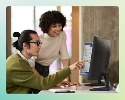 A man wearing headphones and a green sweater pointing at a computer screen while a woman in a white shirt looks on.