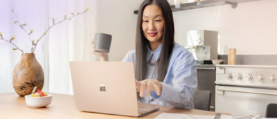 A laptop computer on a table with a cup and a vase with branches nearby.