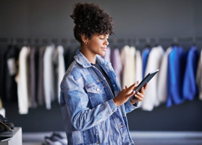 A young woman in a denim jacket using a tablet in a clothing store with racks of clothes in the background.