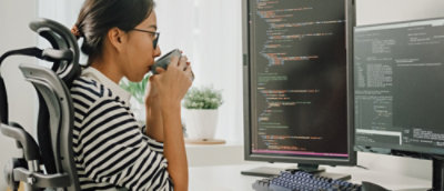 A women sitting in front of computer screen, drinking coffee.