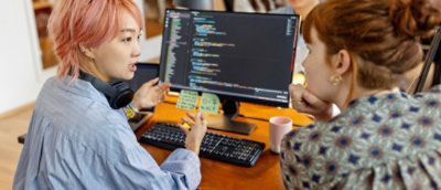 Two women sitting in front of desktop computers and talking.
