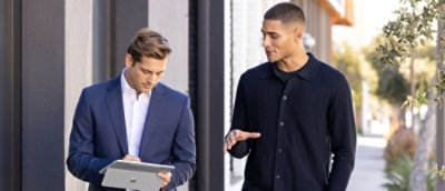 Two men walk outdoors in an urban setting—one in a suit holding a tablet, the other in a dark sweater gesturing thoughtfully.
