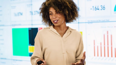 A woman with curly hair presents confidently in front of a digital screen displaying colorful charts and graphs.