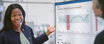 Smiling woman gesturing to a digital screen displaying production data with graphs.