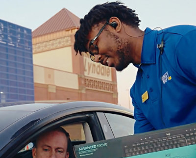 A man wearing glasses is talking to a man sitting inside a car.