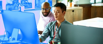 A man with glasses and a beard is seated at a desk, looking at a computer monitor.