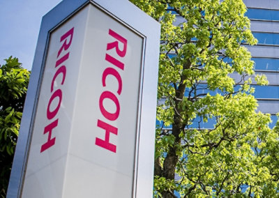 Ricoh sign outside an office building with trees and glass windows in the background.
