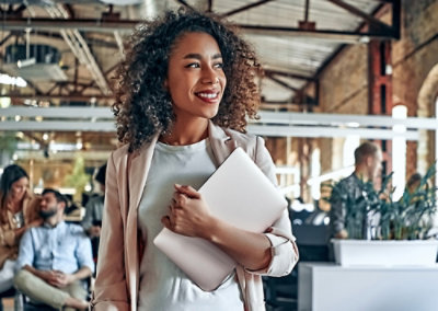 Person holding a laptop and walking through a modern office with coworkers in the background.
