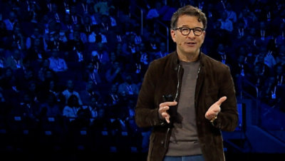A man standing at a podium, in the background multiple pictures are displayed on the screen.