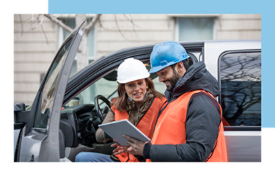 Two people in construction hardhats conversing while using a tablet.