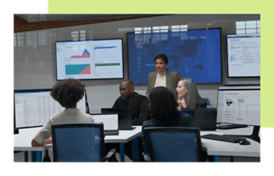 Team collaborating in an office with multiple screens showing data dashboards and a world map, while a woman leads the discussion.