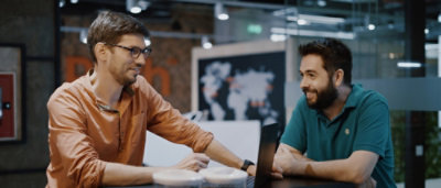 Two men, one in a green shirt and one in an orange shirt, sitting at a table looking at a laptop.